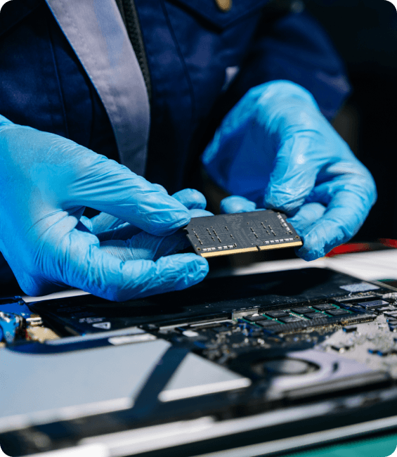 Técnico especializado usando luvas azuis segurando um pente de memória RAM sobre a placa-mãe aberta durante serviços de consertos em notebooks.